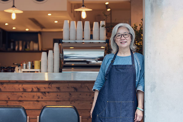 middle aged asian female coffee shop owner standing outside of her open cafe