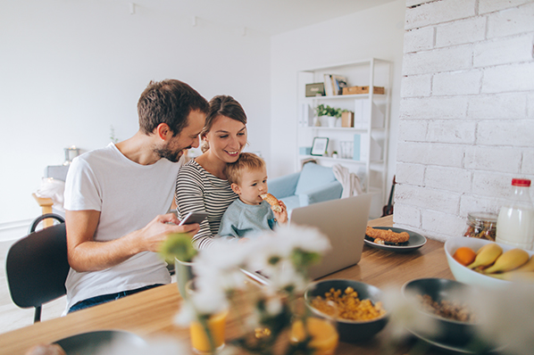 young parents feeding their young child at the kitchen island while looking at the laptop