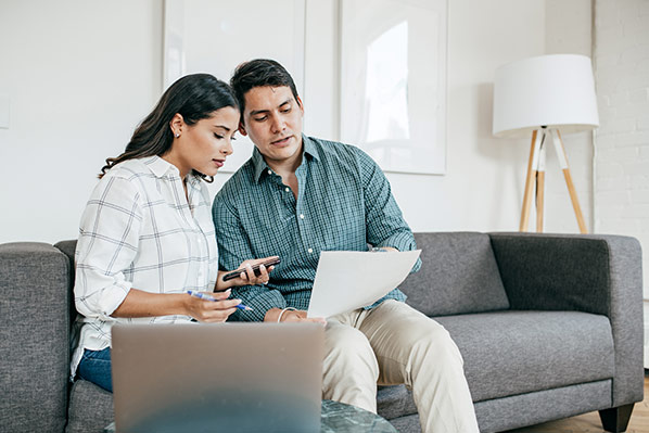 husband and wife sitting on their couch looking at a sheet of paper with pen and a calculator handy