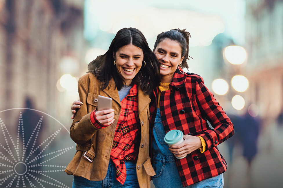 two women laughing walking arm-in-arm holding coffee cups