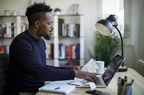 image of a young black male at his laptop with papers in hand and notebook on desk