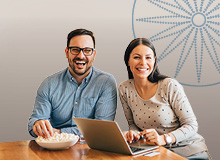 smiling couple sitting at the table looking at their laptop