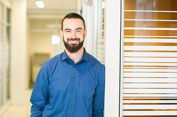 Image of Alex Brocklebank in a blue dress shirt leaning on the office glass wall