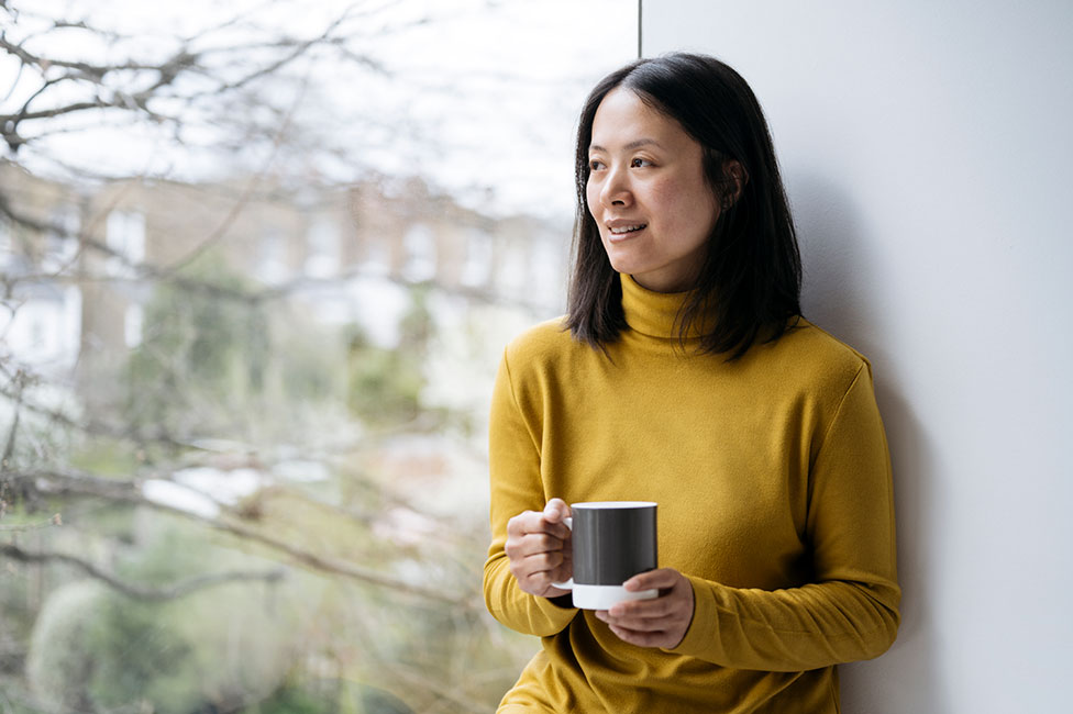 middle-aged Asian woman in a yellow sweater holding a mug and looking out the window