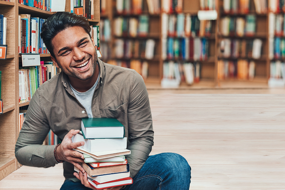 young man smiling and holding a stack of books in the library.png